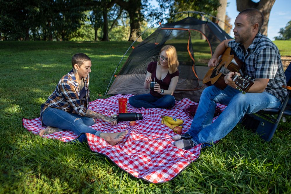 Three friends sit around a picnic blanket near a tent, one playing guitar while the others hold mate gourds.