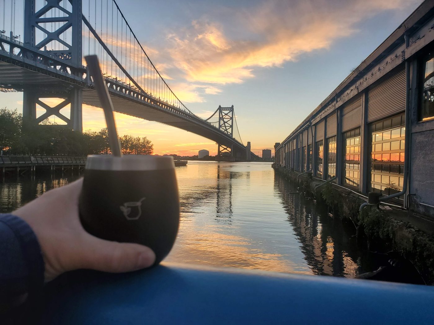 A hand holds a Yerba Crew mate gourd against a sunset view of the Ben Franklin Bridge in Philadelphia.