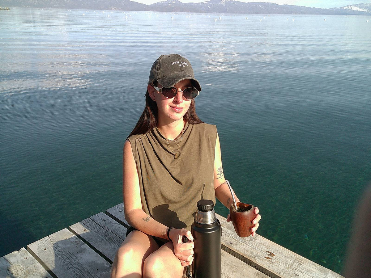 A smiling woman on a lake dock holding a wooden mate gourd with a thermos beside her.