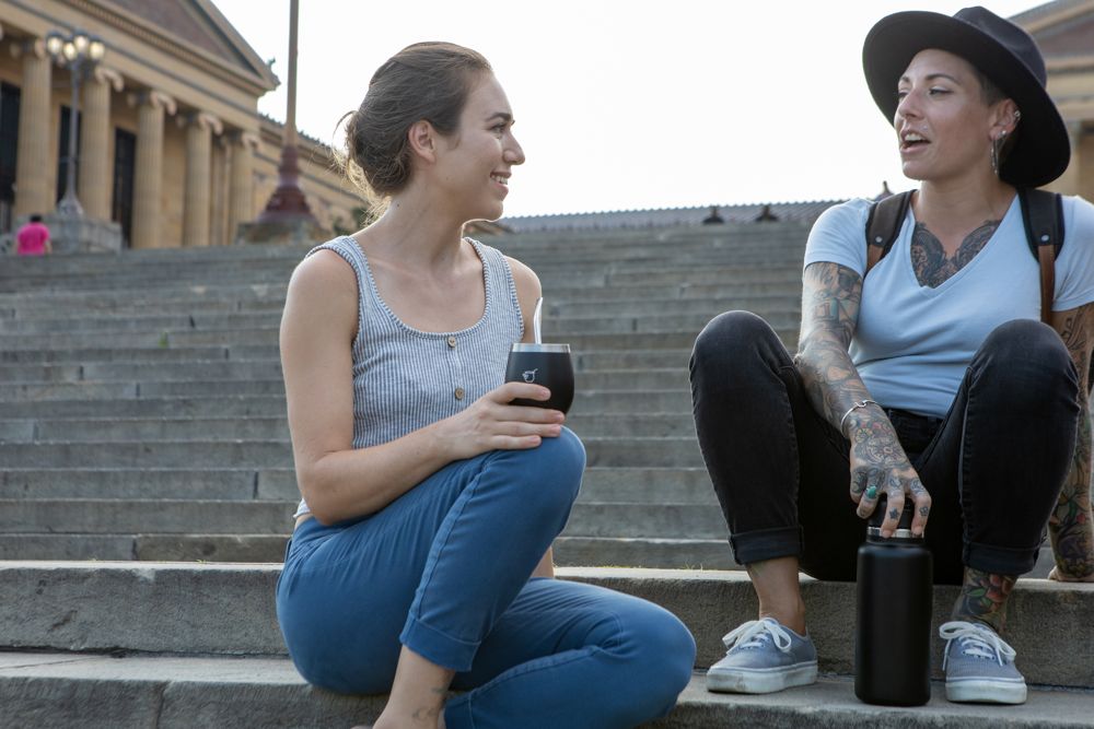 Two women sit and chat on stone museum steps, drinking yerba mate together.