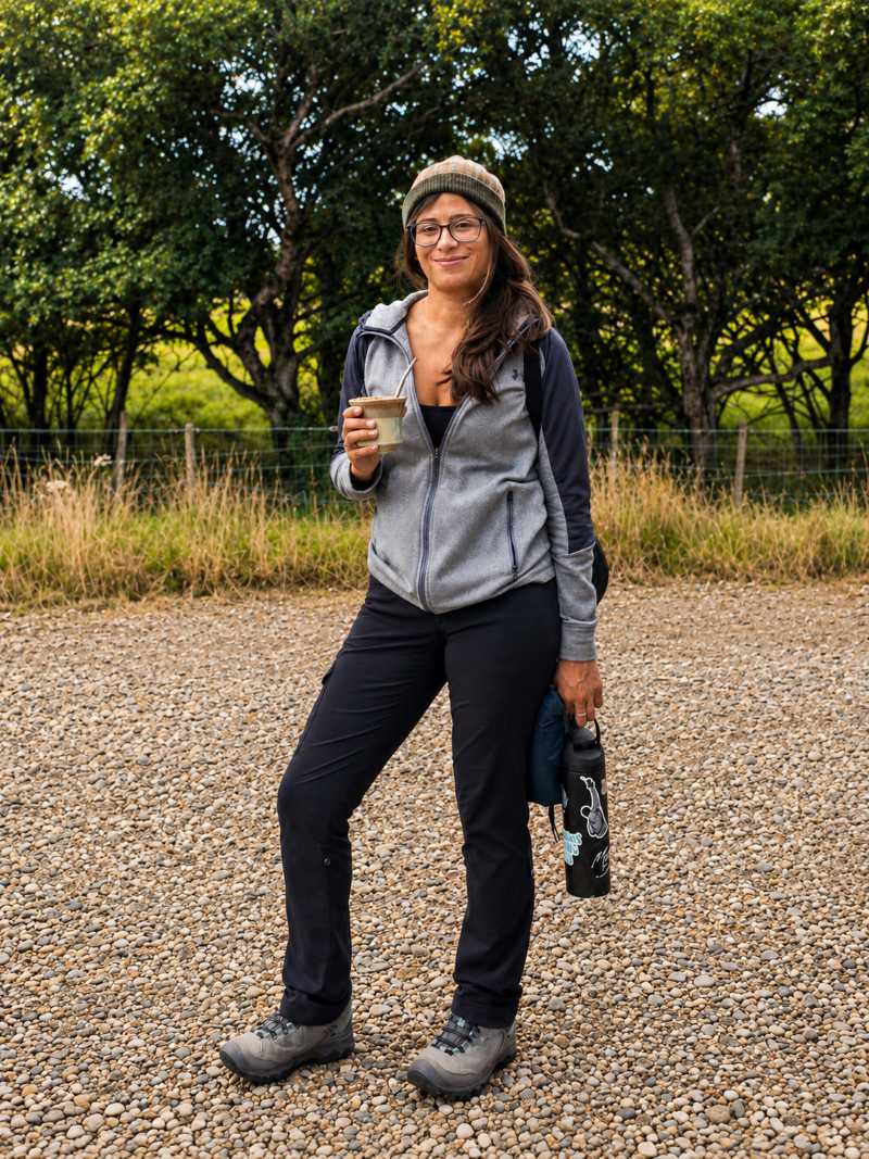 A woman wearing a gray zip-up fleece, black pants, and hiking boots stands on a gravel path outdoors, holding a yerba mate cup and a water bottle. Trees and grass are visible in the background.