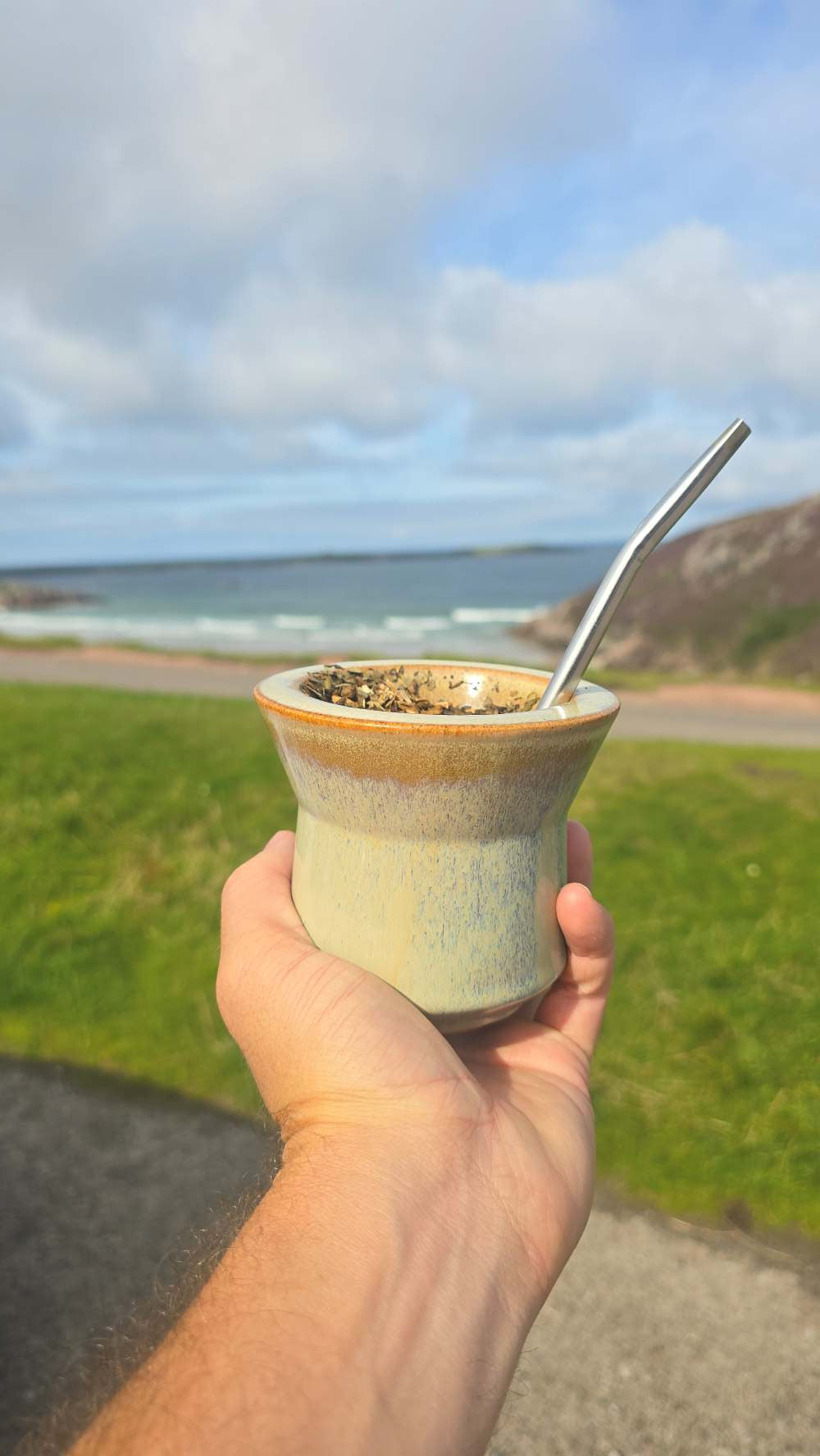 A hand holding a small ceramic mate cup with a metal bombilla, set against a blurred outdoor background of grass, ocean, and cloudy sky.