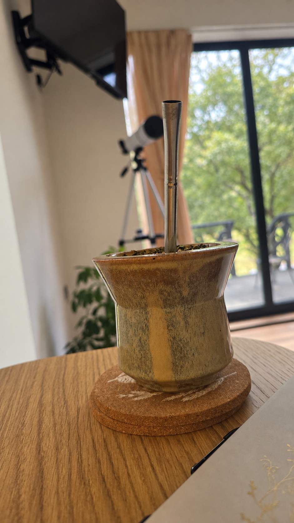 A handmade ceramic mate cup with a wooden base and a silver metal bombilla, set on a wooden table in a home environment with a window and telescope in the background.