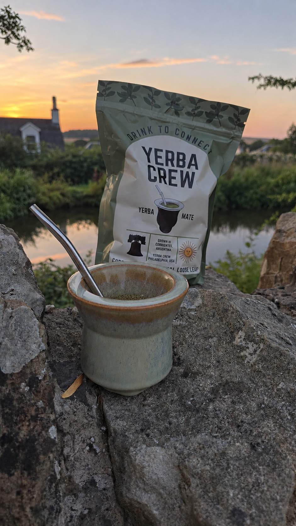 A pouch of Yerba Crew yerba mate sits on a large stone in front of a ceramic mate cup with a spoon, set against a scenic outdoor background with a river and sunset.