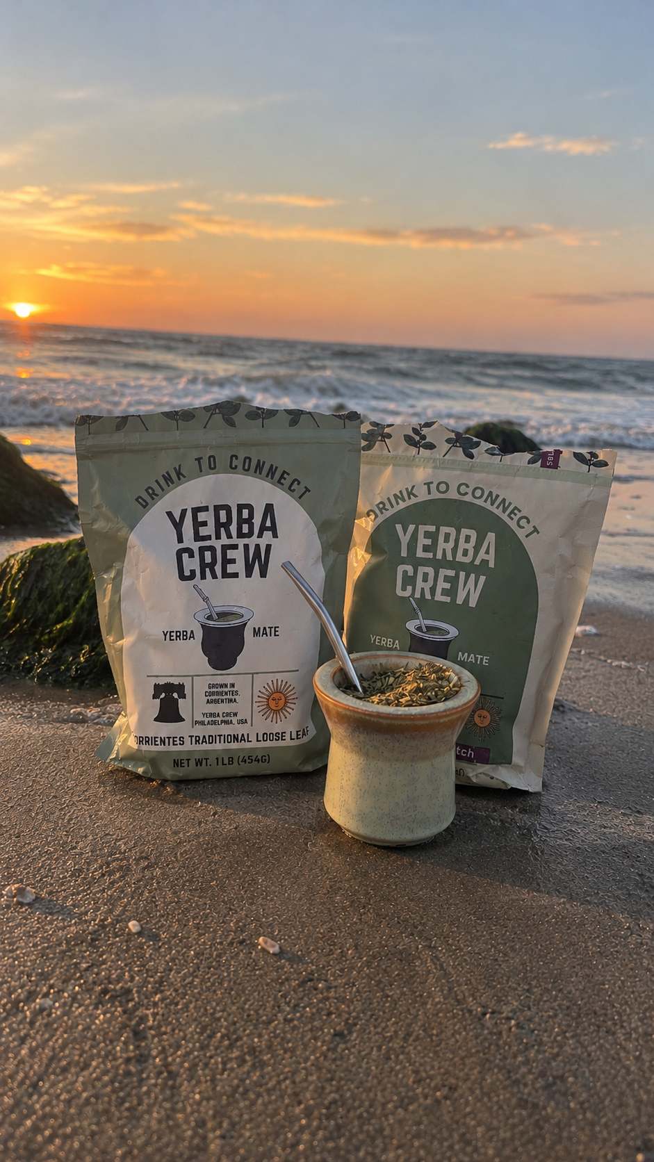 Two bags of Yerba Crew yerba mate tea and a small ceramic cup with a bombilla on a sandy beach at sunset, with the ocean and sky in the background.