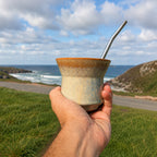 The Iruya ceramic mate gourd with a Bombilla Plana straw, held up overlooking a coastal cliff road in Scotland.