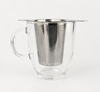 A winged stainless steel infuser basket resting on the rim of a clear glass mug.