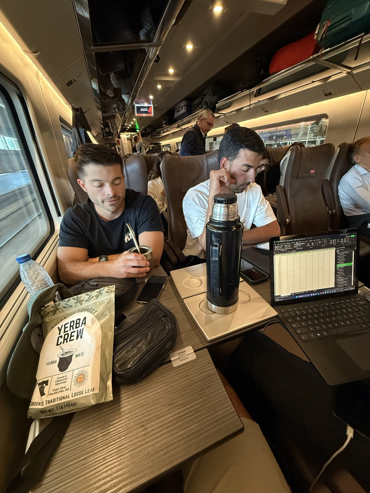 Two men sit on a train with a Yerba Crew bag, thermos and gourd, one working on a laptop.