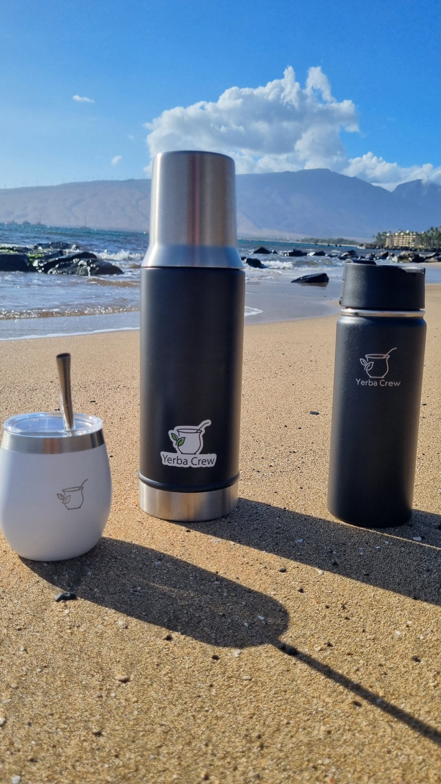 Two Yerba Crew thermoses and a mate tumbler stand on a sandy beach with waves and mountains behind.